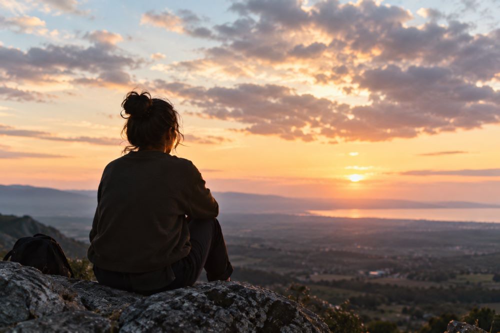 Persona sentada de espaldas sobre unas rocas, contemplando un paisaje montañoso y el mar al atardecer, bajo un cielo cubierto de nubes iluminadas por una luz cálida.