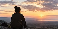 Persona sentada de espaldas sobre unas rocas, contemplando un paisaje montañoso y el mar al atardecer, bajo un cielo cubierto de nubes iluminadas por una luz cálida.