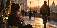 Una mujer escribe en un cuaderno sentada en un banco mientras un hombre camina delante de ella por una calle al atardecer, bajo una cálida luz dorada.