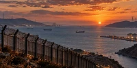 Vista panorámica de un estrecho marítimo al atardecer, con montañas difuminadas, barcos mercantes, infraestructuras energéticas y una valla fronteriza en primer plano, bajo un cielo anaranjado.
