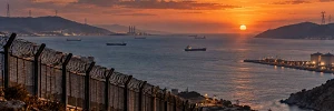 Vista panorámica de un estrecho marítimo al atardecer, con montañas difuminadas, barcos mercantes, infraestructuras energéticas y una valla fronteriza en primer plano, bajo un cielo anaranjado.